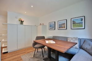 Un salon moderne avec une table à manger en bois, un canapé gris et un tapis gris. Trois tableaux encadrés ornent le mur, ainsi qu'un vase rempli de fleurs et un meuble blanc avec des étagères.