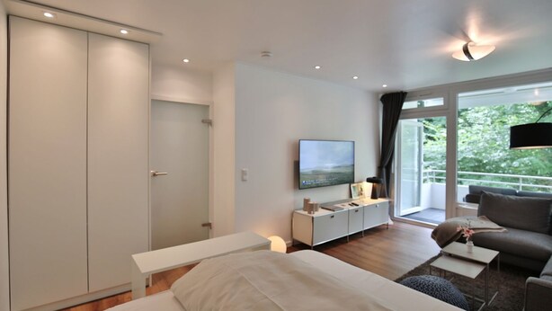 A modern bedroom with a bed, a TV on a white console, a sofa, and a balcony with a view of greenery. - Domizil Strandallee 30 Apartment 8, Casa Bolz - Domizil Strandallee 30 (Timmendorfer Strand)
