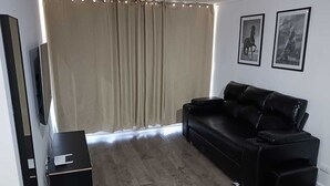 Una sala de estar con un sofá de cuero negro, un televisor de pantalla plana, una pequeña mesita auxiliar negra y un ventanal con cortinas.