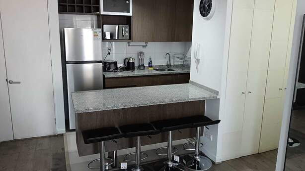 Una cocina moderna con microondas integrado, refrigerador de acero inoxidable y encimera de granito. Hay dos taburetes de bar con patas cromadas.