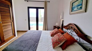A bedroom with a bed, pillows, a nightstand, a lamp, a painting, and a view of the outdoors. - Anna Holiday Home, private heated pool (Montaña Roja)