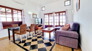 A living room with a checkered floor, a sofa, a dining table with chairs, a TV, and large windows with curtains. - Anna Holiday Home, private heated pool (Montaña Roja)