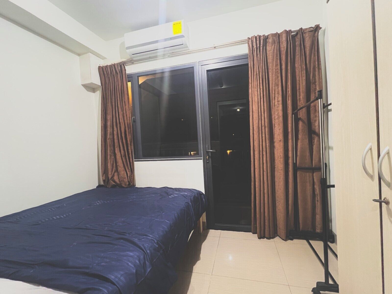 A bedroom with a bed, an air conditioning unit, a window with curtains, and a door.