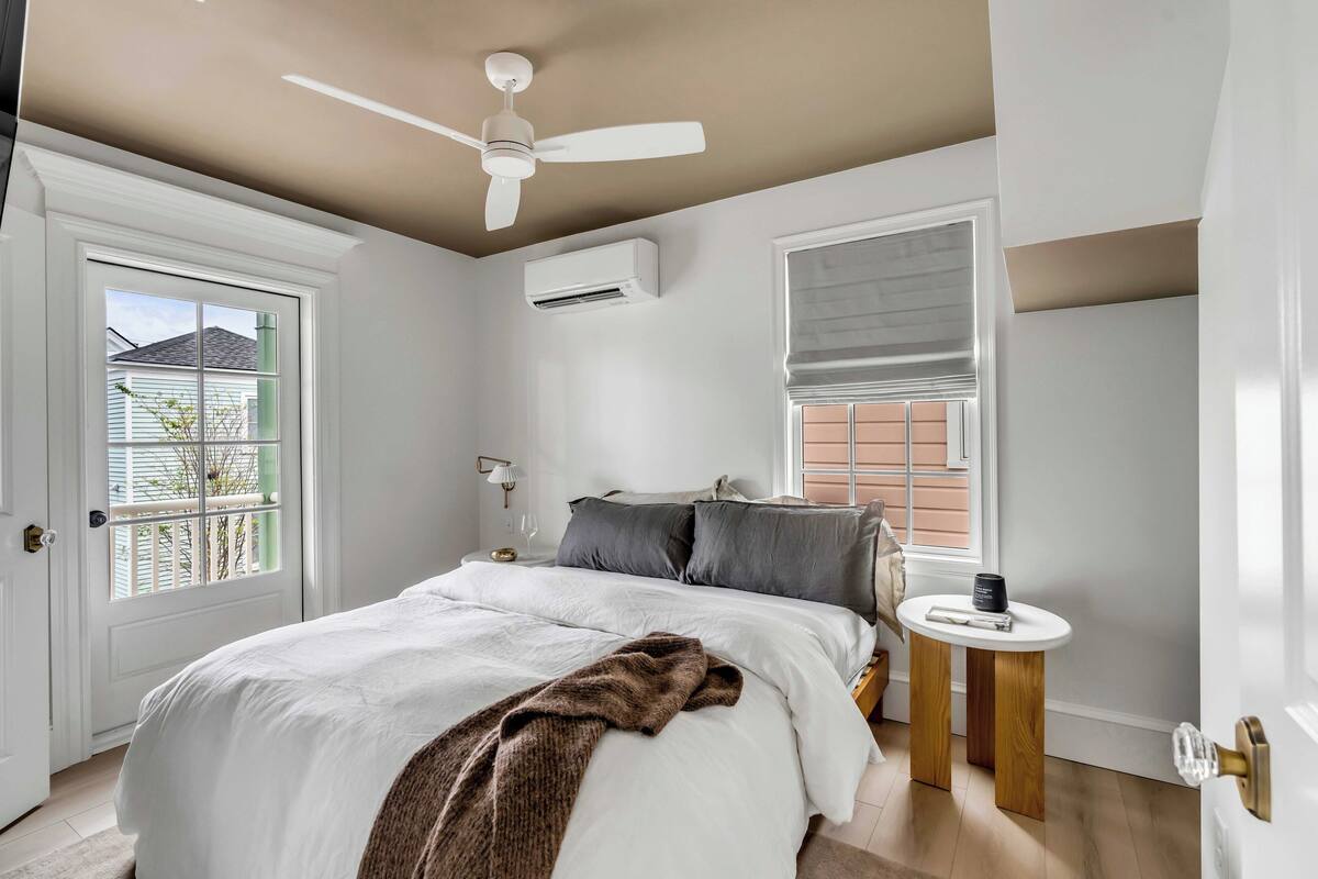 Quarto com cama, ventoinha no teto, ar condicionado, janela com persianas e uma pequena mesa com uma xícara.