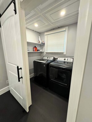 A compact laundry room with a dryer and washing machine, a white door, and a window with blinds. - The Park House  A 5 Bedroom Retreat w/Game Room & (Huntington)