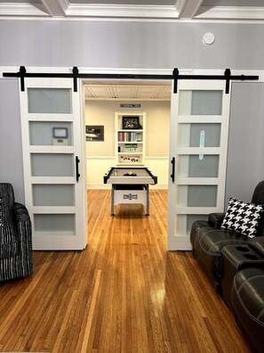 A room with a wooden floor, a foosball table, a sofa, and a couch with a patterned pillow.