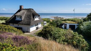 Una casa de techo de paja con fachada blanca, ubicada cerca de una playa con una bandera al fondo.