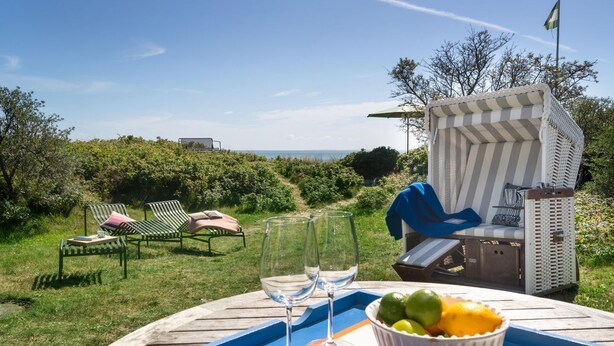 Una mesa con bandeja, vasos y un cuenco de fruta, rodeada de sillones y una silla a rayas, con vistas a un paisaje pintoresco.