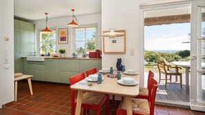 Cocina moderna con armarios verdes, zona de comedor con sillas rojas y vista al océano a través de amplias ventanas.