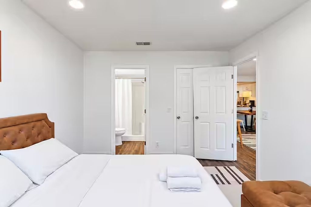 A hotel room with a bed, a bathroom visible through an open door, and a dining area in the background.