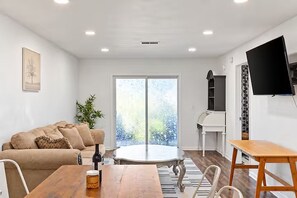 A living room with a sofa, a coffee table, and a TV. There is a large window with a view of trees.