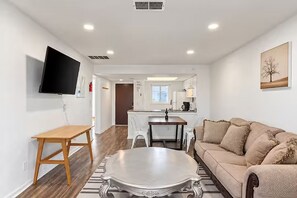 A modern living room with a flat-screen TV, a wooden table, a sofa, and a kitchen in the background.