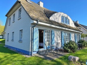 Une maison à deux étages avec un toit en pente, une véranda avec des chaises et un petit jardin.
