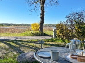 Un espace de restauration en plein air avec une table dressée pour deux personnes, offrant une vue sur une étendue d’eau et un champ verdoyant.