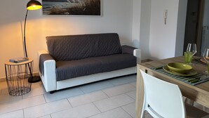 Ein modernes Wohnzimmer mit einem grauen Sofa, einer stehenden Stehlampe, einem Esstisch mit einer grünen Schüssel und einem Bild an der Wand.