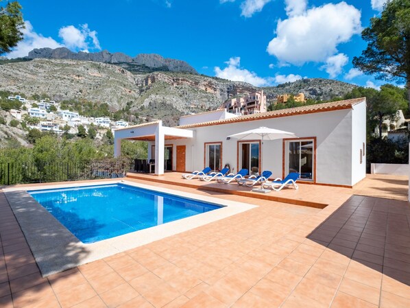 Una villa moderna con piscina, tumbonas y vistas a las colinas circundantes.