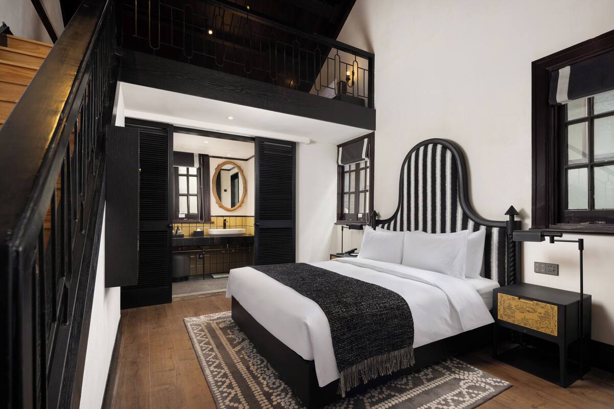 Quarto moderno com esquema de cores preto e branco, uma cama grande, mesa de cabeceira e vista para o banheiro.