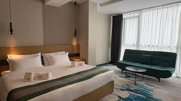 Una habitación de hotel con una cama grande, un sofá verde, una mesita pequeña y una ventana con cortinas.