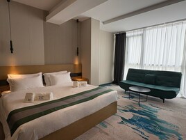 Una habitación de hotel con una cama grande, un sofá verde, una mesita pequeña y una ventana con cortinas.