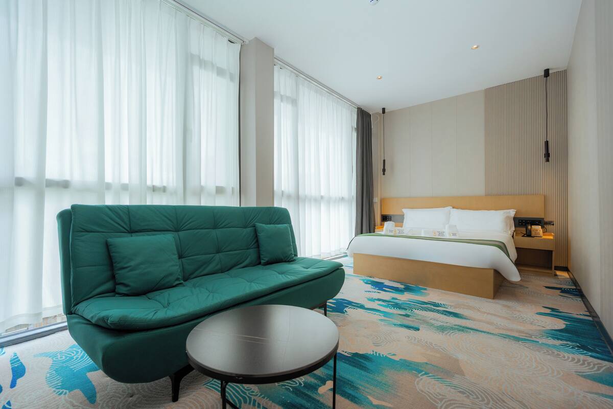 Een hotelkamer met een groene bank, een klein rond tafeltje, een bed met witte lakens en een groot raam met dunne gordijnen.