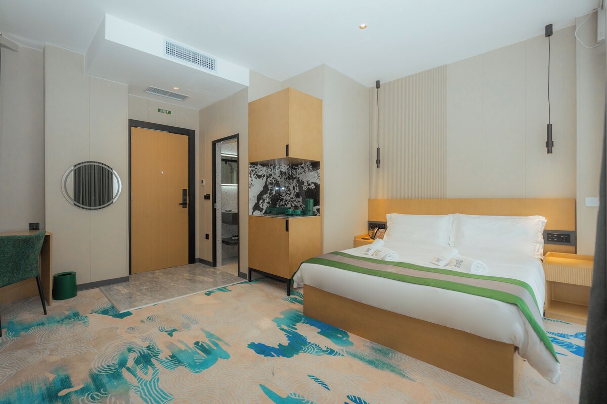 A hotel room with a large bed, a desk, a chair, a trash bin, and a door.