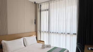 Une chambre d’hôtel avec un grand lit, une tête de lit, une table de chevet, une commode, une fenêtre avec des rideaux légers et une vue sur l’extérieur.