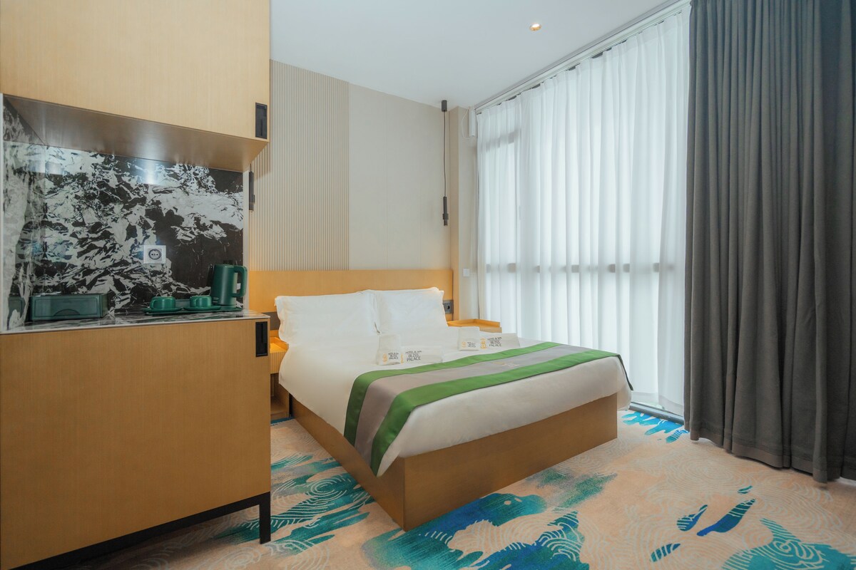 Habitación de hotel con cama, escritorio con una taza verde, ventana con cortinas y alfombra estampada.