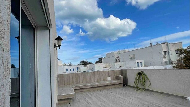 A rooftop terrace with a wooden floor, a glass door, and a view of neighboring buildings under a partly cloudy sky. - Chic Luxury Duplex in The Heart ofla Marsa - Walk to Beach and Cafés (Marsa)