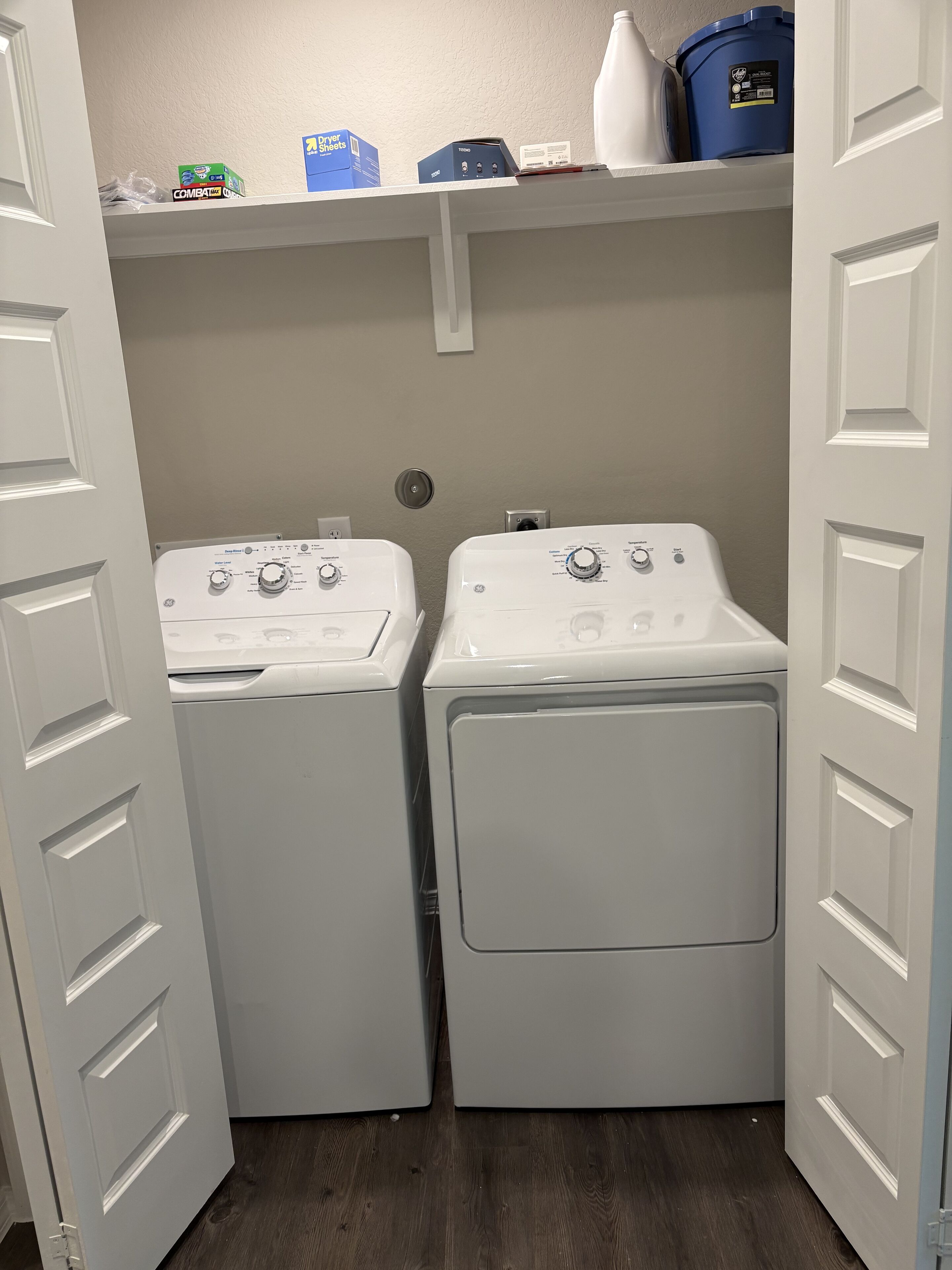 A laundry room with two front-loading washing machines and a shelf above containing cleaning supplies.