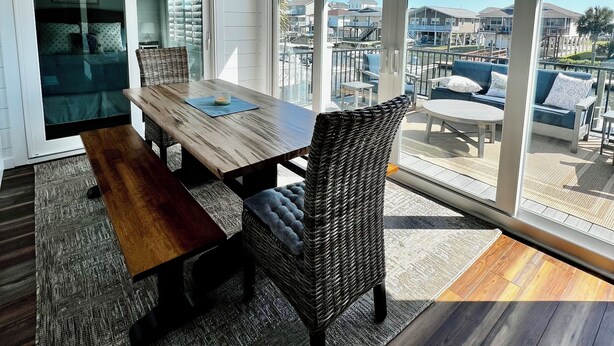 Un comedor con mesa de madera y sillas de mimbre, ventanas amplias con vista a una playa y edificios, y un ventilador de techo.