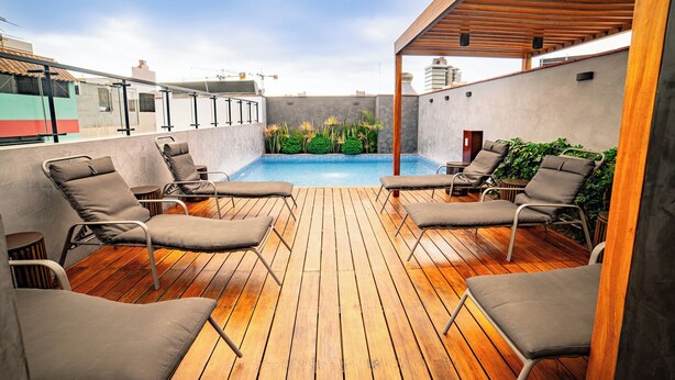 A rooftop pool area with wooden decking, lounge chairs, and a view of the city skyline. - Beautiful and Well Located (Miraflores)