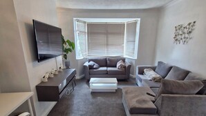 A modern living room with a flat-screen TV, a sofa, a coffee table, and a desk with a chair. - Cosy 3-Bed Home  close to city and shops (Coventry)