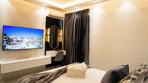 A modern hotel room with a flat-screen TV mounted on the wall, a desk with a chair, a bed with a grey blanket, and a decorative wall hanging.