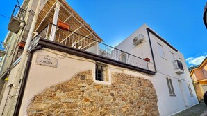 Una stretta via con un edificio bianco caratterizzato da un muro in pietra, un balcone in metallo e un cartello che recita 'Via Nobiltà'.