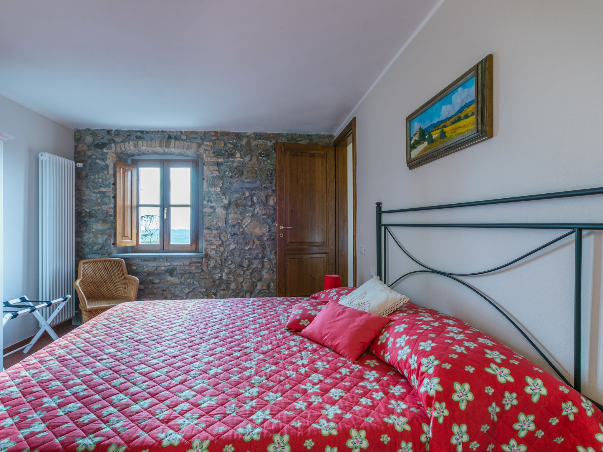 A bedroom with a stone wall, a window, a bed with a floral quilt, a chair, and a painting on the wall.
