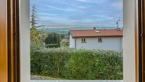 Blick aus einem Fenster auf ein Gebäude mit rotem Ziegeldach, umgeben von Grün und einer entfernten Landschaft.