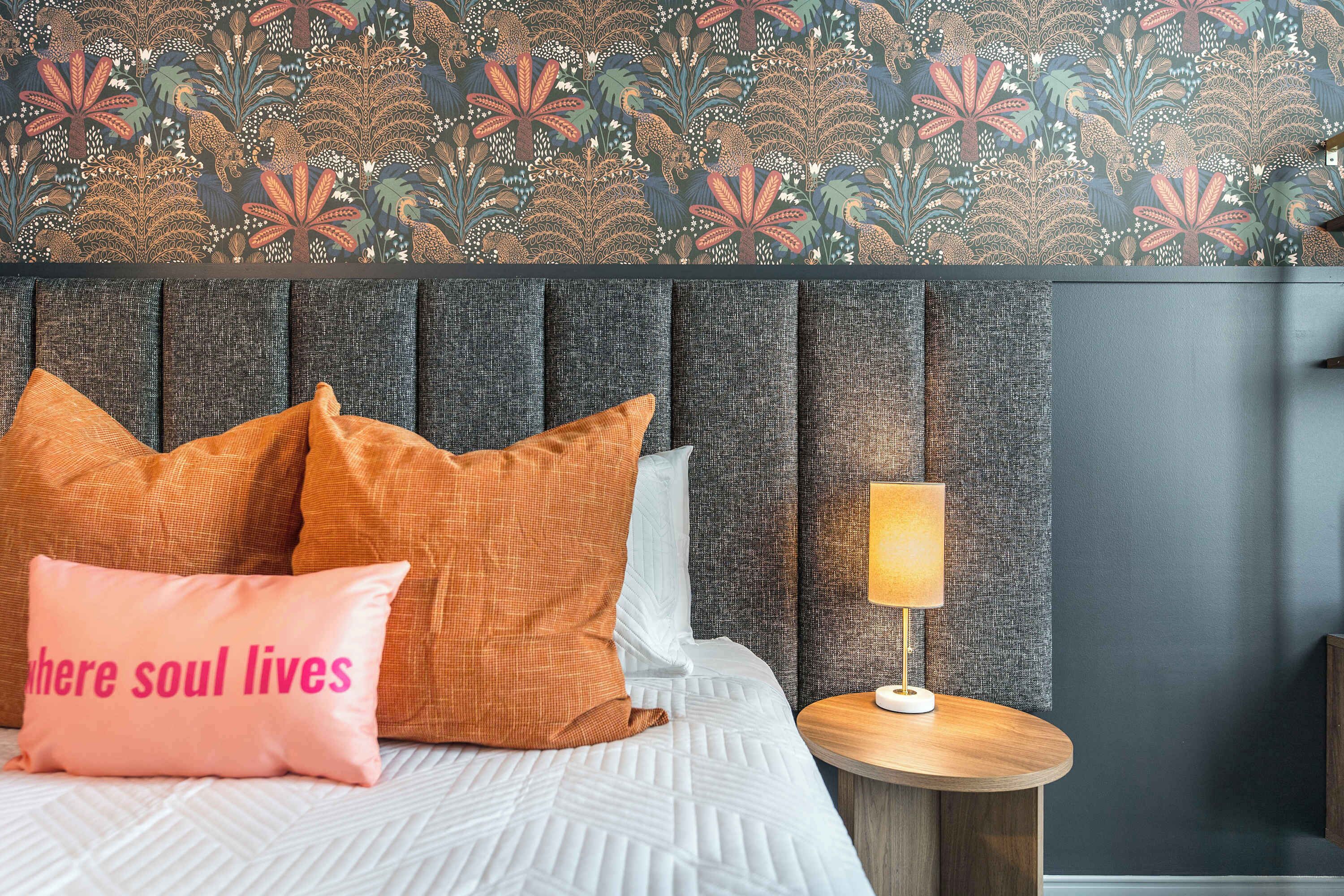 Un lit avec des coussins à motifs, une lampe de chevet et un mur recouvert d’un papier peint à motifs floraux.