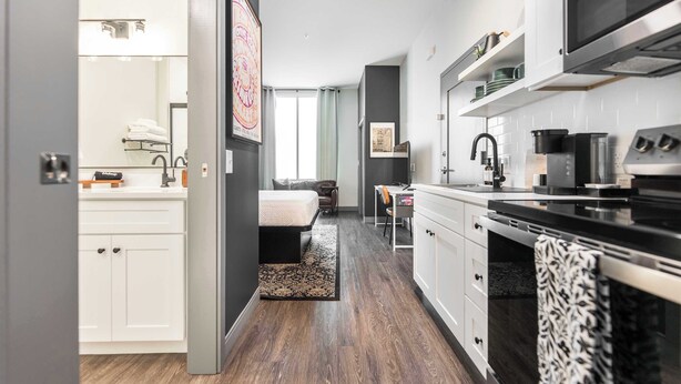 Una cocina moderna con microondas, estufa y armarios, un dormitorio con cama y escritorio, y un baño con lavamanos y toallero.