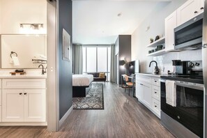 Une cuisine moderne avec des meubles blancs, un four noir et un micro-ondes. On aperçoit un lit avec une tête de lit sombre et un canapé gris au fond.