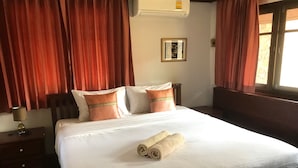 Un lit bien fait, avec du linge de lit blanc et deux oreillers orange, une table de chevet en bois avec une lampe, et une fenêtre avec des rideaux rouges.