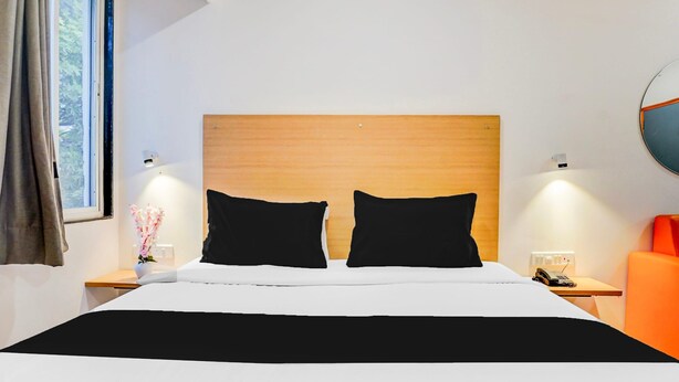 Habitación de hotel con una cama grande, dos almohadas negras, un cabecero de madera, una mesita de noche con una lámpara, un teléfono y una silla naranja.
