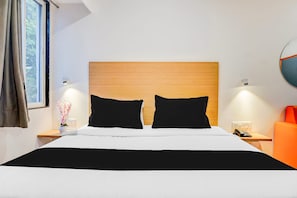 A hotel room with a large bed, two black pillows, a wooden headboard, a nightstand with a lamp, a telephone, and an orange chair. - Elegant Urban Stays at Hadapsar Pune (Pune)