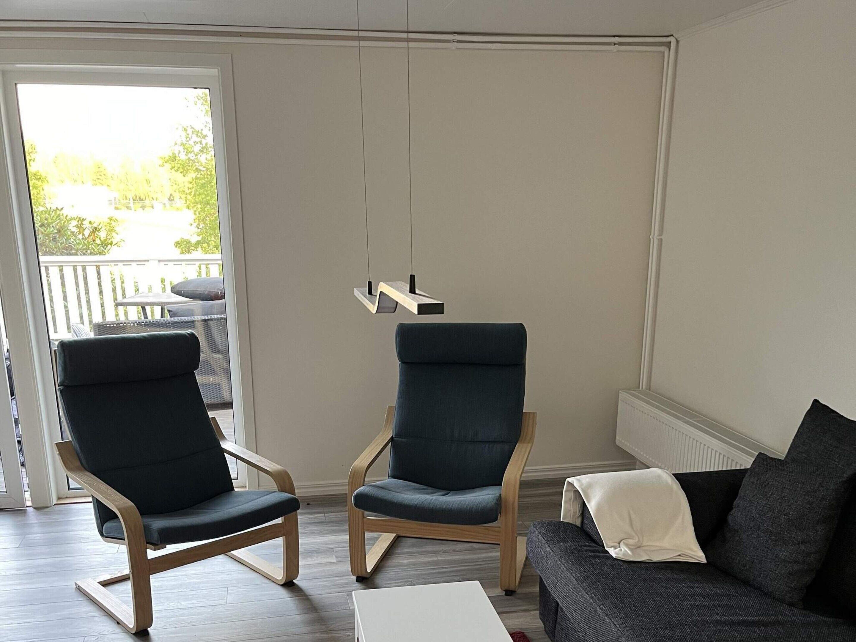 A modern living room with two dark armchairs, a grey sofa, and a white coffee table. A wooden shelf with a hanging light is mounted on the wall.