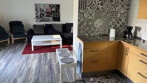Un soggiorno moderno con un divano, un tavolino bianco e un tappeto rosso. È presente un angolo cottura con piastrelle decorate e mobili in legno.