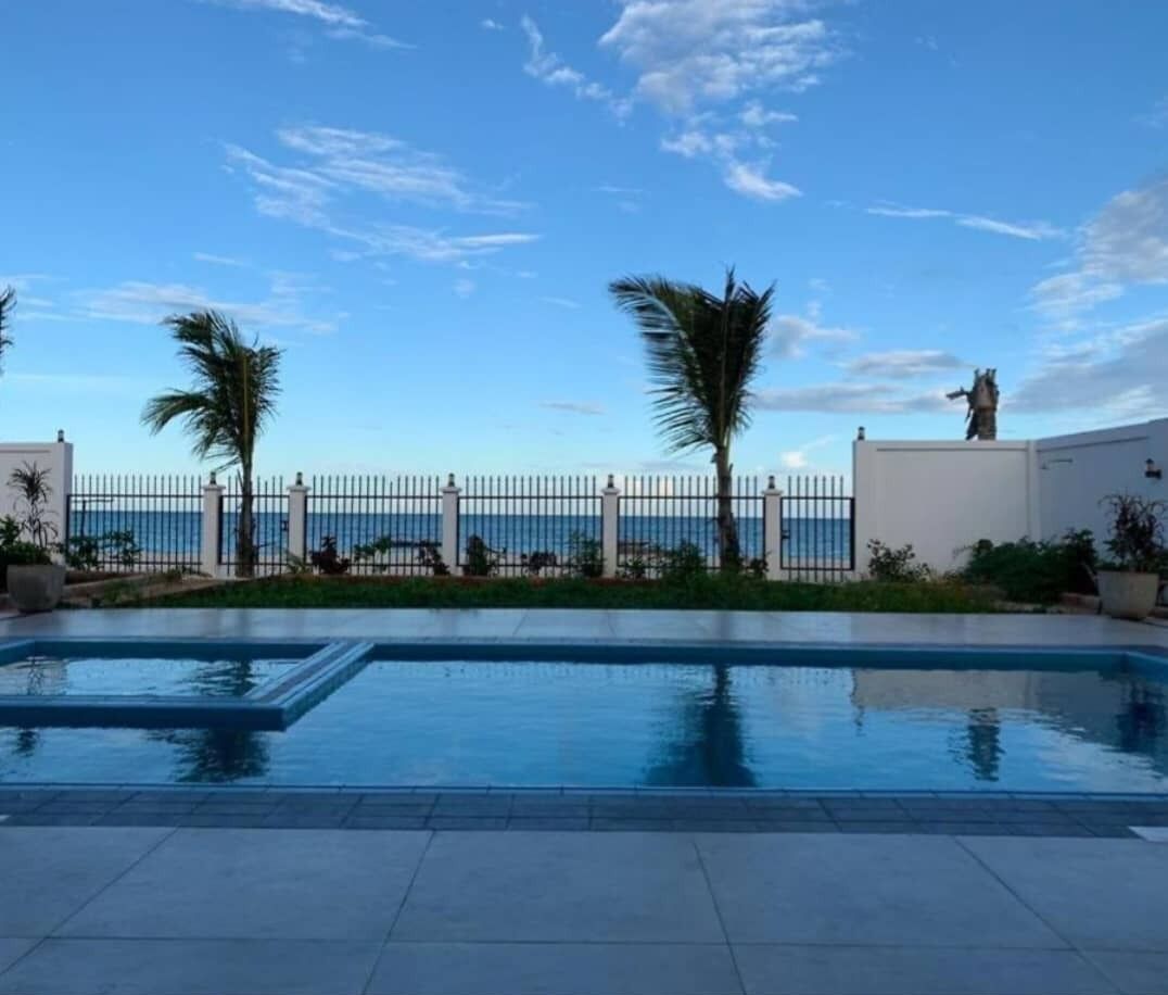 Une piscine avec vue sur la mer, entourée d’une clôture blanche et de palmiers.