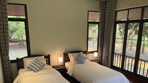 Ein Zimmer mit zwei Betten, Holz-Kopfteilen und Nachttischen mit Lampen. Große Fenster mit Vorhängen lassen natürliches Licht und Blick auf Bäume herein.