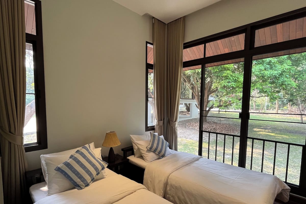 A room with two beds, a lamp, and large windows with a view of greenery outside.