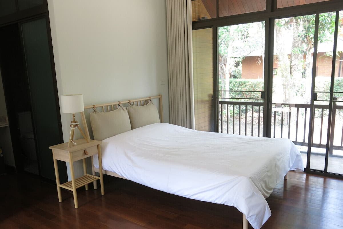 A bedroom with a wooden bed, a bedside table with a lamp, and a view of greenery through large windows.