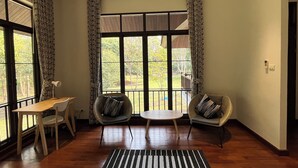 Ein Zimmer mit Holzboden, zwei Sesseln, einem kleinen Tisch und einem großen Fenster mit Vorhängen.
