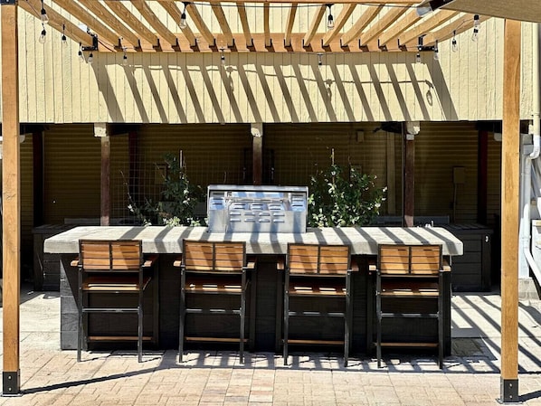 Zona de bar al aire libre con una parrilla, cuatro taburetes de madera y una pérgola.